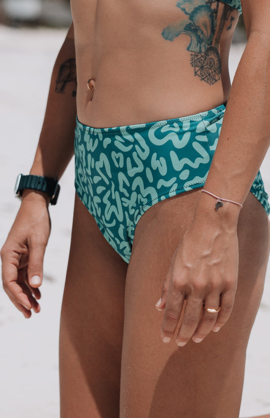 Person wearing teal swim bottoms with white pattern on a beach.